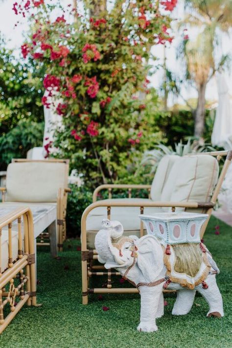 Outdoor seating area with beige cushions and a decorative white elephant stool. Green grass and vibrant red flowers are in the background. Palm Beach Decor Interior Design, West Palm Beach Interior Design, Rattan Accessories, Palm Beach Interior Design, Palm Royale, Beach Decor Living Room, Palm Beach Decor, Pool Vibes, Palm Beach Chic