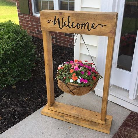 Its that time of the year again for flowers! Display them in a new way by letting us make a hanging basket planter stand with a carving of welcome into the wood! #displayyourstory #homedecor #planterstand #hangingbasket #hangingbasketstand #floralarrangement #cocoliner #homeimprovement #frontdoordecor #palletwooddecor #reclaimedwood #farmhousedecor #outdoordecor #frontporchdecor #welcomesign #carvedwood #diy #diyhomedecor #handpainted #springtime Planter Sign, Welcome Hanging, Diy Hanging Planter, Support Pour Plante, Diy Planter Box, Plant Stands Outdoor, Cozy Fall Decor, Diy Plant Stand, Wood Shop Projects