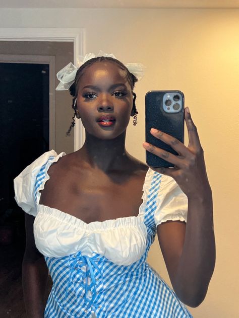 Dark skin black woman taking a photo in the mirror with a Dorothy halloween costume on. She has two space buns with a blue bow on each one. She has red lipstick and has a semi-serious look Dorothy Costume Makeup, Dorothy And Good Witch Costume, Dorothy Gale Makeup, Dorothy Halloween Costume Aesthetic, Dorothy Good Witch Bad Witch Costume, Halloween Costumes Dorothy, Dorothy Makeup, Dorothy Gale Costume, Dorothy Hair