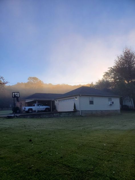 The top half of the photo shows a brilliantly blue, cloudless sky, wisps of fog illuminated by the sun rising over the hilly horizon. The trees in the background are hazy and indistinct. The lower half of the photo features a ranch style house with white siding, looking small in the large green lawn it sits in. It is dark, thrown into sharp contrast with the sky above. Sun Rising, Huntsville Alabama, House Sitting, The Fog, A Hill, Sit Back, A House, Alabama, The Sun