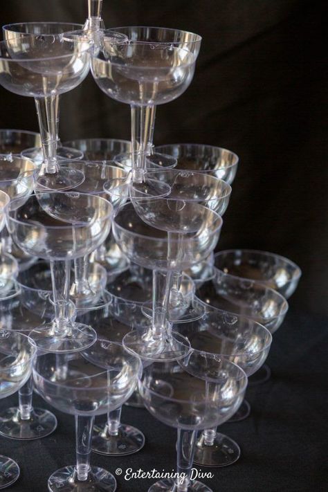 This DIY champagne glass tower is made from plastic coupe glasses that are glued together so it won't fall over or break! It makes a great centerpiece for a Great Gatsby party, roaring 20's event or speakeasy. #entertainingdiva #gatsbyparty #roaring20s #speakeasyparty #centerpiece #diypartydecor Champagne Fountain Diy, How To Make A Champagne Tower, Champagne Tower Diy, Roaring 20s Food, 1930 Party, Champagne Glass Tower, 1920 Wedding, Plastic Champagne Glasses, Glass Tower