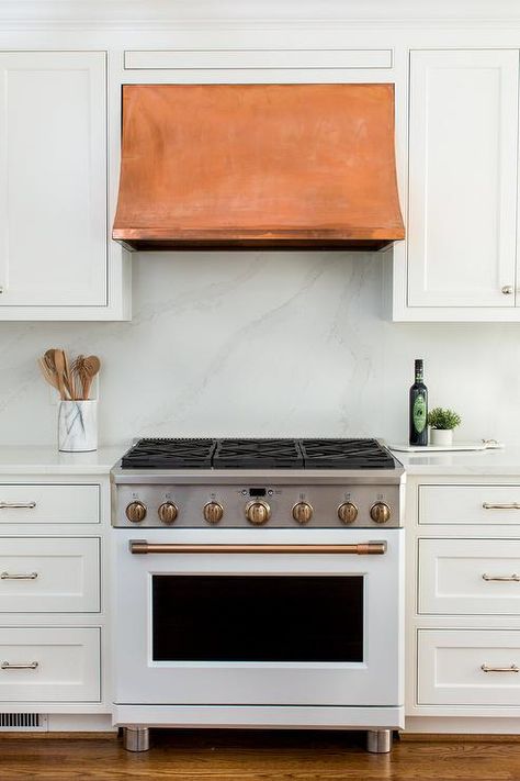 Fixed between white inset cabinets, a copper range hood is mounted against a honed marble backsplash and over a white stove with brass trim flanked by white cabinets topped with a honed marble top. White Kitchen Vent Hood, White Stove, Montgomery Homes, Kitchen Island Tops, Kitchen Vent Hood, Inset Cabinetry, Copper Hood, Kitchen Vent, Inset Cabinets