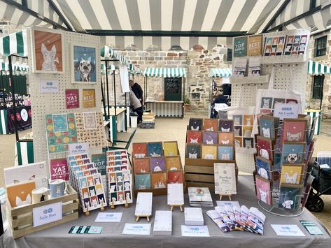 The photograph is landscape, showing a market display stall with two white pin boards one at either back side creating walls. The table is covered in a grey table cloth, and all across it and on the walls are brightly coloured cards, tote bags, prints, notebooks and mugs, with black and white colouring books and memo pads too. Each item has at least one pet illustration on it, from cats & dogs, to rabbits and guinea pigs! Market Stall Display, Stall Display, Brand Colours, Price Signs, House Gardens, Market Displays, Market Stall, Market Stalls, Colour Palettes