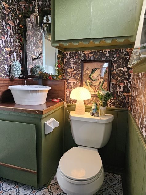 Maximalist bathroom with mossy, green painted cabinets, dark mushroom forest wallpaper, wooden vanity top, vessel sink, mushroom lamp, and frog pictures. Maximalism Cottagecore, Frog Bathroom Ideas, Frog And Mushroom Room Decor, Frog Themed Bathroom, Frog Wallpaper Bathroom, Cottagecore Frog Aesthetic, Frog Bathroom, Cottagecore Frog, Diy Remodeling