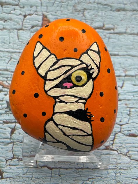 Mummy Cat with Glow in the Dark Eye, Halloween Art, Painted Rock, Stone Painting Measures approximately:  Height 2.75" Length 2" Depth .75" ✽ My painted rocks are natural creek rocks collected locally in middle Tennessee and hand painted by me.  ✽ Your stones are handpicked and hand painted by me with love! Each is a one-of-a-kind piece of art! I hope they bring much joy, happiness, and a smile to you.   ✽ These are natural creek rocks so each stone is unique and no two rocks are identical.  The size, shape, and texture may vary slightly. ✽ Each stone design is drawn by hand, painted with acrylic paints, sealed, and packaged with care to send to your home from my home in middle Tennessee.   ✽ All my creations are sealed to protect and preserve the quality. ✽ Want a custom design?  If you d Fall Rock Art Ideas, Fun Halloween Art Projects, Mummy Painted Rocks, Painted Halloween Rocks, Cat Painted Rocks, Halloween Rock Painting, Halloween Painted Rocks, Painted Skulls, Stone Paintings