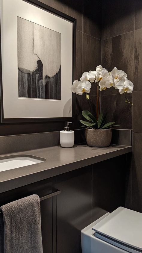 Modern bathroom with dark brown walls, white toilet, gray countertop, framed artwork, towel rack, and orchid vase. Bathroom Dark Countertop, Small Bathroom Solutions, Brown Countertop, Bathroom Dark, Dark Brown Walls, Modern Small Bathrooms, Bathroom Inspiration Modern, Grey Countertops, Contemporary Color Palette