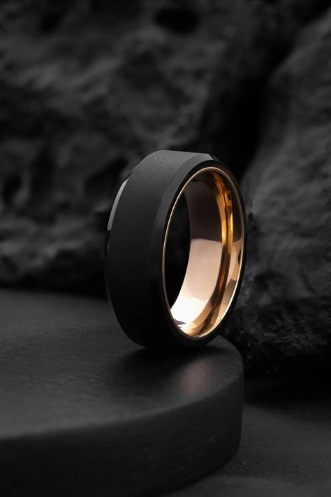 Inspired by Thanatos, the god of peaceful eternity, this Thanatos Wedding Band is a symbol of timeless love and strength. Combining minimalist design with exceptional durability, this ring features a rugged hammered black tungsten carbide exterior, polished to a refined finish with diamond paste. The bold texture of the black tungsten is complemented by a warm rose gold interior and an offset inlay, creating a unique and elegant contrast that symbolizes a balance between resilience and beauty. B Black Wedding Bands For Men, Black Mens Ring, Modern Mens Wedding Bands, Rose Gold Interior, Black Wedding Bands, Engagement Ring Modern, Black Tungsten Carbide Ring, Modern Wedding Band, Wedding Bands For Men