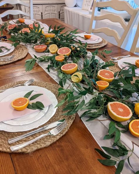 If you're looking for a centerpiece for a beautiful dinner party, this aesthetic might be the one for you. In the middle of the table, place a soft white table runner and decorate it with fresh green leaves accompanied by oranges, lemons, and grapefruit that pair with the natural wood table. Finally, place round rattan placemats below white plates and place a single half-orange and a sprig of greenery on each plate. Lime Table Decor, Orange And Green Table Decor, Clementine Centerpiece, Spanish Table Setting, Mediterranean Feast, Centerpiece For Dining Room Table, Lemon Centerpiece, Red White And Green Christmas, Plate Centerpiece