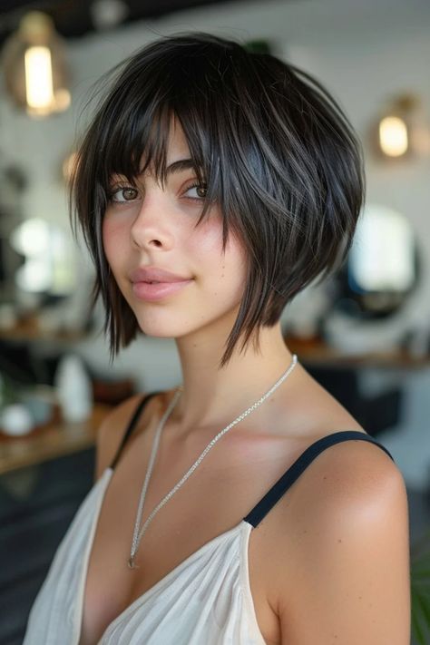 Woman with a short, dark bob hairstyle and bangs, wearing a white top and necklace, smiling slightly in a cozy indoor setting. Shaggy Angled Bob With Bangs, Inverted Bob With Bangs Medium, A Line Stacked Bob Haircut, Angled Shaggy Bob, Short Angled Bob With Bangs, Angled Short Bob, Angled Bobs With Bangs, Straight Angled Bob, Short Aline Bob