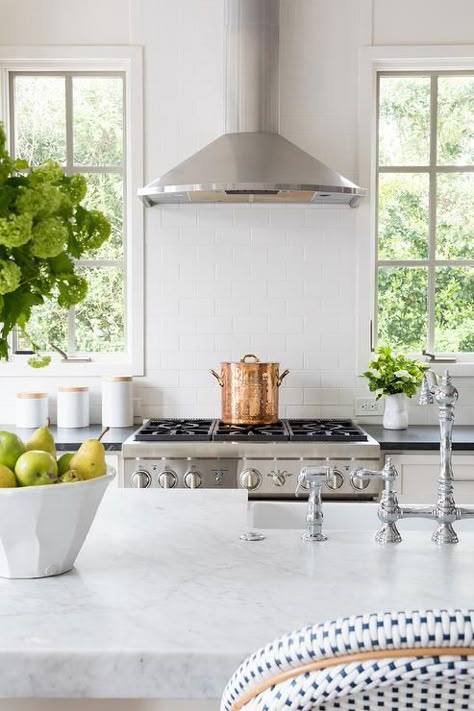 A stainless steel range hood is fixed between windows to a white subway tiled cooktop over a stainless steel oven range flanked by white cabinets contrasted with a black countertop. Range Between Windows, Kitchen Window Above Sink, Window Above Sink, Birch Interior, Flat Front Cabinets, No Upper Cabinets, Beautiful White Kitchens, Floating Kitchen Shelves, Kitchen Cooktop