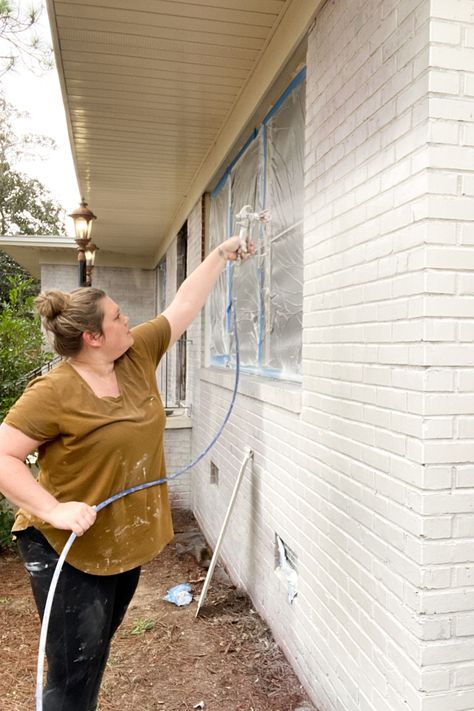 Woman using Graco Magnum 262800 X5 Stand Airless Paint Sprayer to paint exterior brick home white with paint sprayer. Instructions of how to paint a brick house with pros and cons. Blue painters tape with sheeting covering windows. Painted White Brick House, Painting Brick Exterior, Red Brick Paint, White Brick Exterior, Brick Exterior Makeover, Brick Painted White, House Exterior Before And After, Painted Brick House Exterior, Red Brick House Exterior