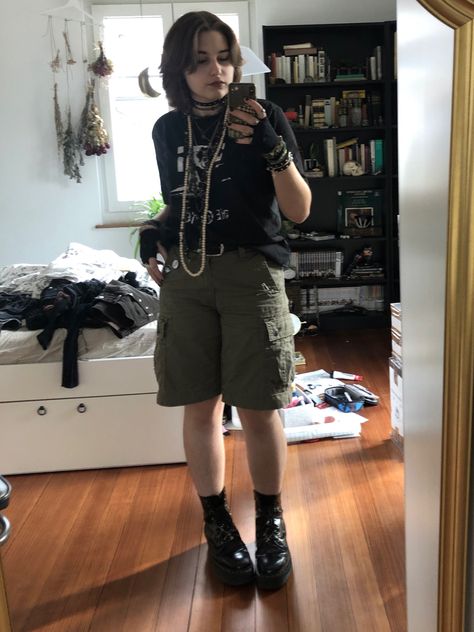 A mid-sized person/girl wearing a    black the cure shirt, green cargo shorts, fingerless gloves, platform dr. Martens and multiple dark green and gray crystal rings and bracelets. 		The person is also wearing multiple silver necklaces. Summer Goth Masc, Alt Button Up Shirt Outfit, Eco Goth Fashion, Eco Punk Fashion, Masc Skirt Outfit, Lillie Core, Masc Fairy Grunge Outfit, Masc Summer Outfits For Women, Adrogonus Outfits