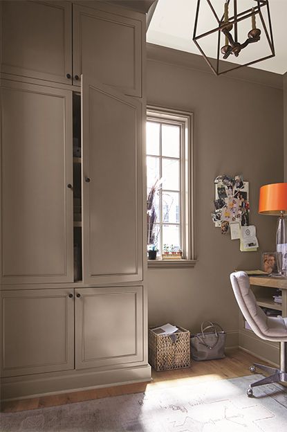 A taupe-painted home office with cupboard, beige chair, wooden desk, note board, white ceiling, and abstract chandelier. Best Dark Beige Paint Color, Dark Beige Kitchen Walls, Dellwood Sand Benjamin Moore, Rich Taupe Paint Color, Built In Bookcase Color Ideas, Falcon Brown Benjamin Moore, Taupe Walls And Trim, Taupe Wall Paint Colors, Taupe Dressing Room