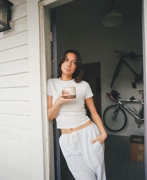 A woman with a cup of coffee leans against the door frame to outside in pajamas Door Frame Poses, Woman Lounging, Leaning Against Door Frame Pose, Lazy Day Photoshoot, Playful Relaxed Fit Sleepwear For Lounging, Coffee Run, Leaning Pose, Casual Ribbed Sleepwear With Relaxed Fit, Cozy Relaxed Fit Summer Sleepwear