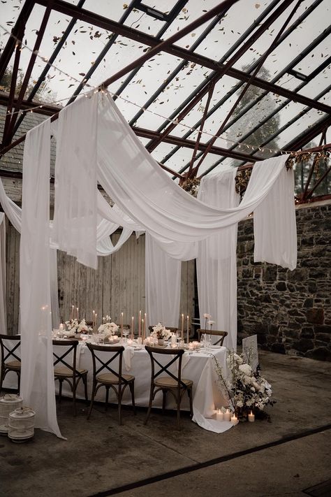 Minimalistic, neutral wedding reception room with glass roof, white wedding drapes, tapered candles and white rose and foliage decorations | Sam Sparks Photography | <a href="https://www.rockmywedding.co.uk/ceiling-drapes-decoration">See more of this real wedding</a> Drape Wedding Reception, Ivory Polyester Tablecloth Wedding, Fabric Drapes Wedding, Candle Table Wedding Decor, Glass Room Wedding, Roof Draping Ideas, Curtains Wedding Decor, Fairy Lights On Ceiling Wedding, Glass Roof Wedding