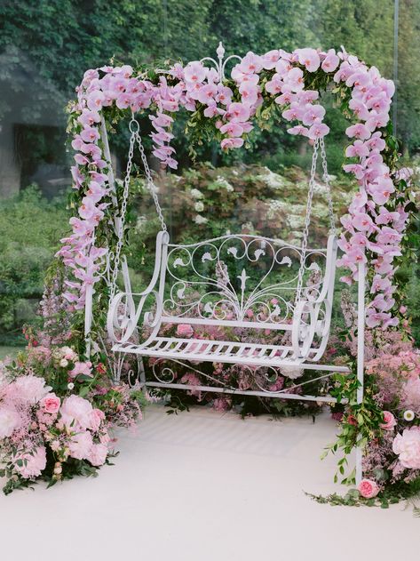 A white swing decorated with cascading pink orchids, standing in front of a green backdrop. Swing Decoration With Flowers, Wrought Iron Garden Furniture, Balcony Herb Gardens, Flower Chair, Wall Partition, Porch Swing Bed, Selfie Wall, Glamorous Decor, Backyard Swings