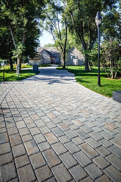 This driveway design is inspired by our Pure paver. Pure brings the larger cobblestone look to permeable pavements for residential and commercial driveways and patios. This interlocking permeable pavement was designed to allow for the proper percolation of surface water into the ground; reducing the risk and severity of flooding, eliminating puddles and speeding up the melting process of snow. Check out our website to shop the look! Side Of Driveway Landscaping, Grey Driveway, Driveway Pavers Design, Driveway Landscaping Ideas, Entrance Landscaping, Driveway Entrance Landscaping, Cobblestone Driveway, Permeable Pavers, Driveway Lighting