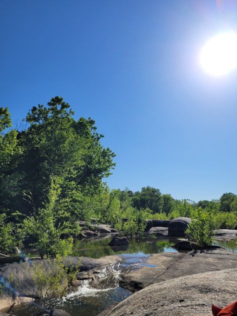 View from sitting on the rocks in the James River. The rocks are large and flat and smooth. Easy enough to walk from rock to rock around the river. The water is shallow and around the rocks. There is movement in the water but not much. It's a sunny day and it's may, so the trees and grass are green. River Aesthetic, Summer Vision, James River, Belle Isle, The James, March 2024, Richmond Va, Summer 2023, Fun Things