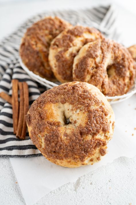 A bowl of cinnamon crunch bagels on the counter next to cinnamon sticks. Homemade Cinnamon Raisin Bagels, Sourdough Cinnamon Crunch Bagels, Cinnamon Streusel Bagels, Cinnamon Sugar Bagels, Cinnamon Bagel Recipe, Cinnamon Crunch Bagel Recipe, Cinnamon Crunch Bagel, Cinnamon Bagel, Panera Bread Copycat