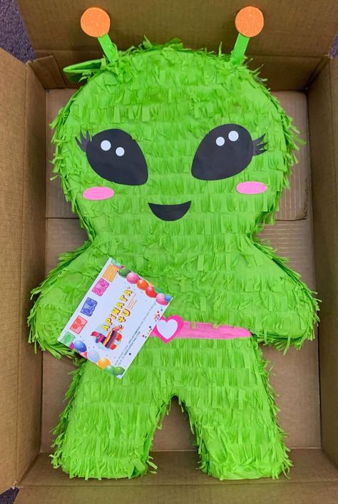 Inside a cardboard box, a green alien piñata with black eyes, pink cheeks, and orange antennas is displayed. The piñata has a pink belt with a heart-shaped buckle, and a branded tag from "apinata4u" is attached. Alien Birthday Party, Alien Birthday, Mini Alcohol Bottles, Pinata Stick, Alien Party, World Party, Kids Party Themes, Birthday Party 21, Birthday Surprise Party