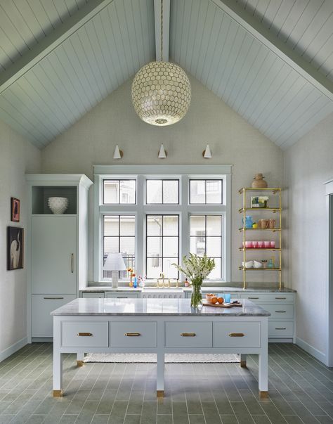 a pale green painted cabinet is used in this outdoor, poolhouse kitchen. The island has drawers and open legs at the bottom. There is a brass shelf holding colorful acrylic cups and glassware for entertaining. There is a large window overlooking the yard, and a very large orb-shaped light fixture of capiz shells and brass. Poolhouse Guesthouse Interiors, Pool House Decor Interior Design, Pool House Kitchenette, Poolhouse Kitchen, Poolhouse Guesthouse, Pool House Interior, Pool House Kitchen, Tub Room, Pool House Bathroom