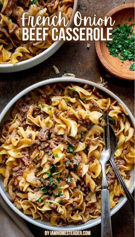 A beige bowl is filled with French onion beef casserole and has a silver spoon and fork in it. Around the bowl is a small wooden bowl of parsley, and another identical bowl of French onion beef casserole, all on a dark countertop. Creamy French Onion Beef And Noodles, French Onion And Beef Noodles, French Onion Beef And Noodles Casserole, French Onion Beef Noodle Bake 12 Tomatoes, French Onion Soup Hamburger Recipes, What To Make With French Onion Soup, Ground Beef And French Onion Soup, Ground Beef Onion Recipes, Cabbage And Noodles With Ground Beef