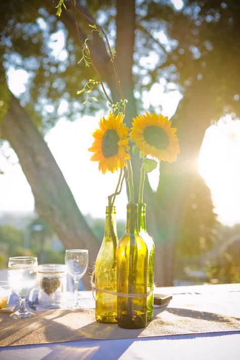 Sunflower and wine bottle centerpieces tied with twine on burlap runner #sunflowers #rusticwedding Sunflower Wedding Vases, Sunflower Bud Vase Centerpiece, Burlap And Sunflower Table Decorations, Wine Centerpiece, Sunflowers On Glass Bottles, Sunflower Centerpieces Wedding Budget-bride.shop, Yellow Wedding Decorations, Wine Theme Wedding, Anniversary Centerpieces