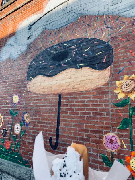 a single donut is pictured in front of a brick wall mural; the donut is a vanilla cream stick with chocolate sprinkles. the mural is filled with flowers that are decorated with cinnamon rolls and donuts inside of them. The middle of the mural features a giant chocolate donut being used as an “umbrella” to block a rain shower of sprinkles. Selfie Wall For Bakery, Bakery Mural, Business Painting, Mural Inspiration, Selfie Wall, Restaurant Themes, Mural Wall Art, Cake Shop, Photo Op