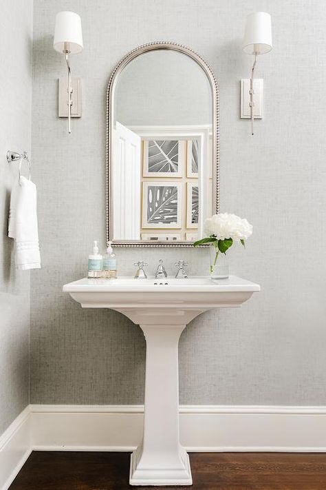 Between nickel sconces mounted to a wall covered in gray wallpaper, an arched beaded mirror hangs over a white pedestal sink finished with a polished nickel cross handle faucet. Modern Bathroom Pedestal Sink, Powder Room Ideas With Wood Floors, Small Powder Bathroom Ideas Pedestal Sink, Mirror For Pedestal Sink, Pedestal Sink Mirror, Half Bathroom Pedestal Sink, Mirror Over Pedestal Sink, Powder Room With Pedestal Sink, Powder Room Layout