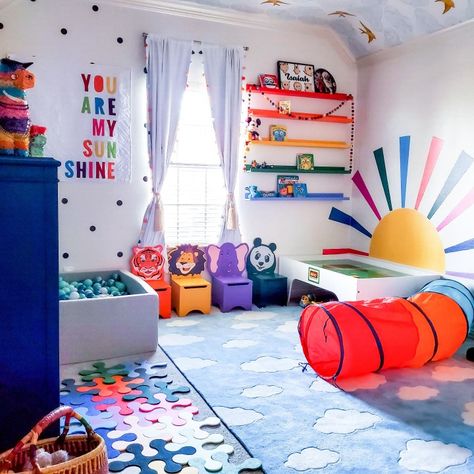 Bright blur rug with white fluffy clouds in center of room. Rainbow color shelves on a wall. Large horizon rising sun with rainbow colored beams. Large ball pit filled with an array of blue hued balls. Black and white polka dots on wall Playroom Ideas Colors, Nursery Decor Bright Colors, Playroom Ideas Rainbow, Bright Color Playroom, Bright Rainbow Bedroom, Playroom Bright Colors, Playroom Decor Colorful, Fun Colorful Nursery Ideas, Colorful Toy Room