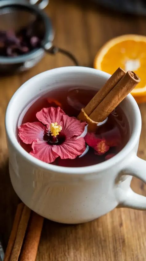 A photo of a warm cup of Hibiscus-Cinnamon tea with a cinnamon stick and orange peel. The tea has a deep red color and is served in a white mug. There are hibiscus flowers and a cinnamon stick in a small strainer at the bottom of the mug. The background is a wooden table. Hibiscus Flower Drink Recipes, Hibiscus Tea Blend Recipe, Rose Water Recipes, Hibiscus Tea Aesthetic, Alkaline Drinks, Ginger Cinnamon Tea, Hibiscus Tea Recipe, Hibiscus Flower Tea, Hibiscus Drink