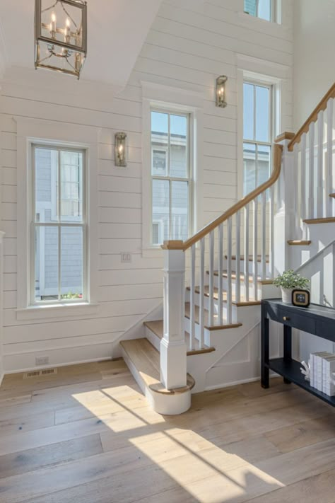 A foyer with white paneled walls, warm light stained wood floors, a winding staircase, beautiful gold sconces and a chandelier, and lots of windows! Coastal House Inside, Stairs Beach House, Coastal Banisters And Railings, Coastal Railings For Stairs, Coastal House Foyer, Coastal Home Staircase, Beach Front House Interior, Coastal Themed House, Beach Home Decor Coastal Style Interior Design