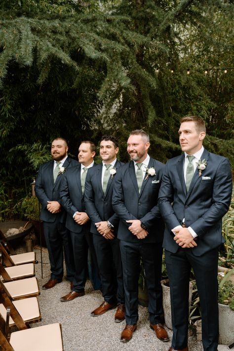 Groomsmen standing watching bride walk down the aisle Navy Blue Suit Green Tie Wedding, Dark Navy Blue Groomsmen Suits, Groomsmen Attire Dark Blue, Deep Blue Groomsmen Suits, Bestman Suit Groomsmen, Eucalyptus Wedding Groomsmen, Black Suits Brown Shoes, Navy Suit With Green Tie, Navy Groomsmen Suits And Bridesmaids