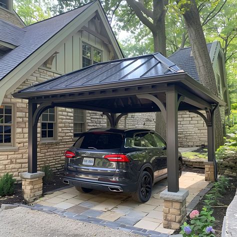 Elegant carport featuring a dark gray metal roof, beautifully complemented by tan stone accents and limestone piers. This open-sided structure shelters a black Volkswagen Tiguan, set against an old cottage-style house with black windows. The landscaping boasts lush greenery, colorful flowers, and two large decorative brown blocks enhancing the aesthetic. Carport Beside House, Beautiful Carport Ideas, Carport Driveway Ideas, Pergola As Carport, Cottage Carport Ideas, Covered Car Port Ideas, Detached Carport Ideas Breezeway, Adding A Carport To Front Of House, Carport With Privacy Wall