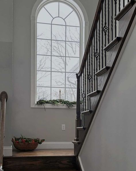 Dark wooden steps let down to a landing that features soft gray walls and a gorgeous arched window styled for the holidays with a garland and simple candle holder. The trim of the window is a classic white that matches the grilles. Staircase Wall Design Modern, Stairwell Window, Modern Stairway, Staircase Wall Design, Stair Bookshelf, Staircase Window, Stairway Wall, Bookshelf Modern, Stairs Window