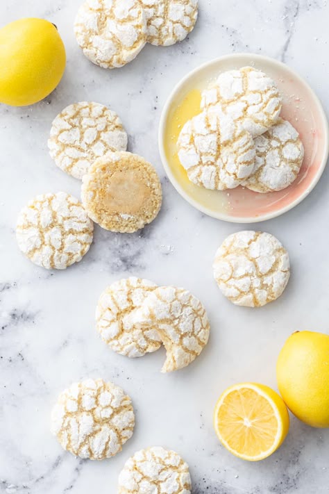 Overhead, scattered Chewy Lemon Almond Crinkle Cookies on a marble surface with a few fresh lemons and three cookies on a small ceramic plate. Lemon Turmeric Crinkle Cookies Nyt, Lemon Powder Cookies, Lemon Amaretti Cookies, Lemon Turmeric Crinkle Cookies, Lemon Tumeric Cookies, Meyer Lemon Cookies, Almond Flour Lemon Cookies, Soft Amaretti Cookies, Egg White Cookies