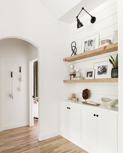 This living room boasts warm wood floors and a black lamp hanging on a white shiplap wall above long wooden floating shelves holding a group of pictures with white mats in beige frames. An off-white bowl is placed on the white cabinets with black knobs underneath the shelves. White Floating Shelves Above Sideboard, California Casual Style, Warm Wood Flooring, Long Floating Shelves, White Shiplap Wall, Wooden Floating Shelves, White Shiplap, California Casual, Building A New Home