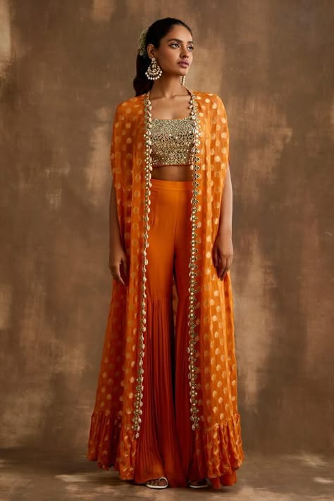 Turmeric orange cape with mirrorwork embellishments and floral patterns. Comes with padded blouse and gharara.
Components: 3
Pattern: Embroidered
Type Of Work: Mirrorwork
Neckline: Cape: Open, Blouse: Square
Sleeve Type: Cape: Cape sleeves, Blouse: Sleeveless
Fabric: Georgette, Lining: Shantoon
Color: Orange
Other Details: 
Attached lining
Model Height: 5ft 9inches wearing size XS
Closure: Blouse: Back drawstring
Occasion: Sangeet,Mehendi and Haldi - Aza Fashions Orange Indian Lehenga, Orange Indo Western Outfits, Cape Indian Outfit, Indian Cape Outfits, Orange Traditional Outfits, Indo Western Sangeet Outfit For Women, Haldi Indowestern Outfit, Orange Haldi Outfit, Indian Haldi Outfit