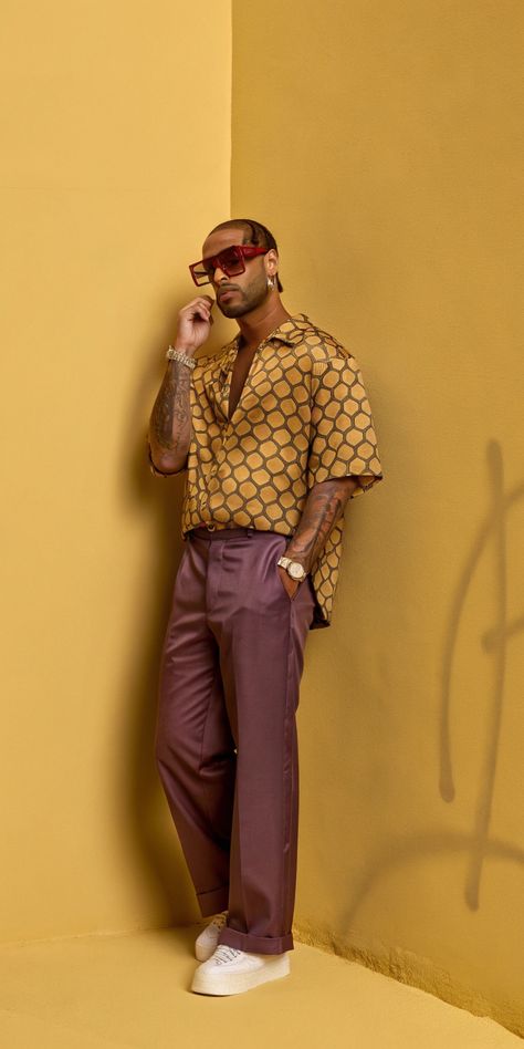 A man wearing a mustard geometric print shirt, purple tailored trousers, red sunglasses, and white sneakers, posing against a mustard yellow wall, showcasing a bold and confident style. Relaxed Tailoring, Vibrant Fashion, Streetwear Chic, Confident Style, Geometric Prints, Bold Accessories, Rich Color Palette, Autumn Outfits, Bold Prints