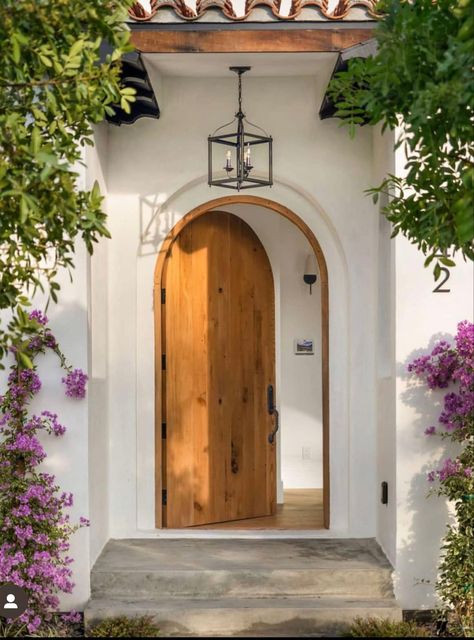 White Stucco exterior, arched wood door, framed by beautiful purple and green vines Spanish Bungalow Exterior, Arched Front Door, Celebrity Home, Celebrity Mansions, Spanish Bungalow, Wood Entry Doors, Bungalow Exterior, Exterior House Color, Arched Doors