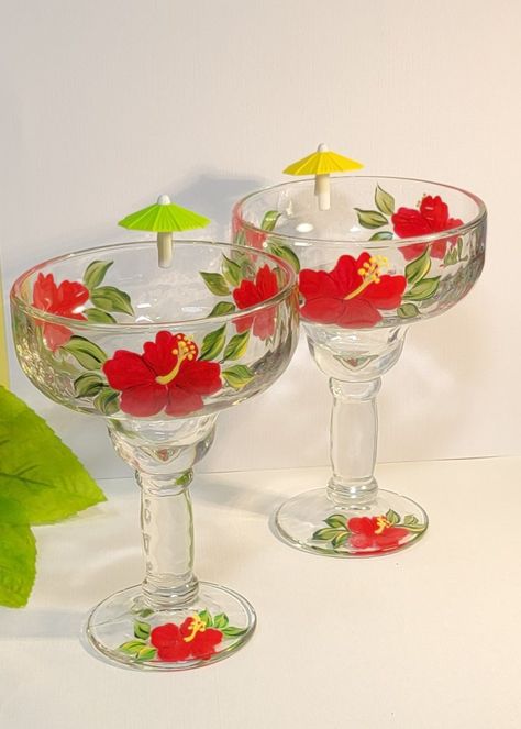 This set includes two hand-painted margarita glasses adorned with red color hibiscus flowers. Each glass showcases three flowers encircling the bowl and a single flower on the base, complemented by green foliage. **While supply lasts, you will receive a complimentary umbrella wine glass marker for each glass ordered. These glasses are made of heavy glass. Measurements:  7.50 inches in height, 4.5 inches in width at the top, and have a generous capacity of 15 ounces. In addition to magenta-colored hibiscus flowers, we also offer varieties in purple, red, and yellow. High-quality glass enamel paint is applied, and the glass undergoes heat treatment to guarantee the durability of the paint design. Although these glasses are dishwasher safe on the top rack, hand washing is recommended to avoid Hibiscus Margarita, Wedding Drinkware, Fiesta Decor, Wine Glass Markers, Glass Showcase, Red Hibiscus, Three Flowers, Margarita Glasses, Fiesta Decorations