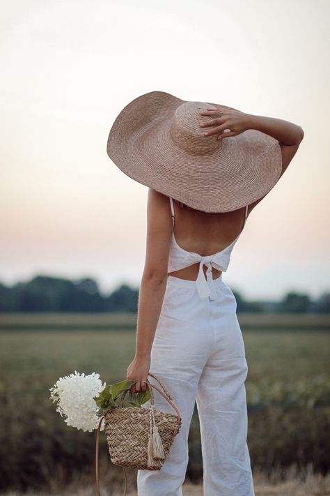 Annie B. standing in field watching sunset in oversized straw hat and white linen jumpsuit taken from behind Outfit With Straw Hat, Large Straw Hat, Poses With A Hat, Holding Hat Pose, Woman From Behind, Swimsuit Shoot, White Linen Jumpsuit, Watching Sunset, Open Dress