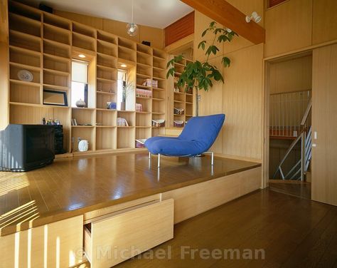 The living room of a low-cost Japanese home in Chiba, Japan, by architect Shintaro Hanazawa. The sitting area is on a raised wooden platform that provides additional storage space, with a grid of wooden shelving covering one wall. Raised Wooden Platform Living Room, Raised Platform Living Room Floors, Platform Living Room Raised, Raised Floor Storage, Raised Floor Living Room, Raised Living Room Floor, Japanese Reading Room, Japan Tiny Apartment, Raised Platform Living Room