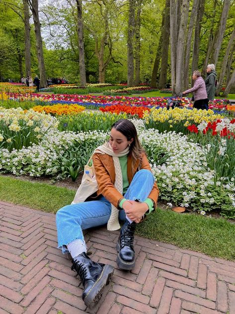 A girl wearing a bright lime green sweater underneath a caramel brown leather trench coat, light blue jeans, and platform black doc martins is sitting crossed-legged next to beds of multicolored tulips. Tulip Garden Outfit Ideas, Keukenhof Gardens Outfit, Amsterdam Tulip Fields Outfit, Netherlands Outfits Spring, Spring Outfits Amsterdam, Keukenhof Outfit, Keukenhof Photoshoot, Amsterdam Outfits Spring, Amsterdam Fashion Spring