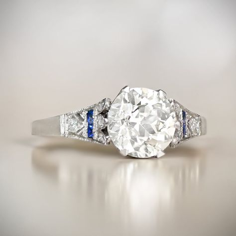 This engagement ring features a 1.60 carat old European cut diamond, J color and VS1 clarity. The geometric shoulders are adorned with French cut sapphire accents and old European cut diamonds. Additional old European cut diamonds are set along the crown-style under-gallery. The total weight of the sapphires is approximately 0.12 carats. The weight of the diamonds on the shoulders and under-gallery is approximately 0.24 carats combined. This ring is handcrafted in platinum and decorated with fin Platinum Engraved Engagement Ring, Antique Diamond And Sapphire Engagement Ring, Saphire Engament Ring Set Silver, 25 Anniversary Rings, Shiney Things, Blue Diamond Wedding Ring, Antique Engagement Rings Sapphire, European Cut Diamond Engagement Ring, Diamond Sapphire Ring