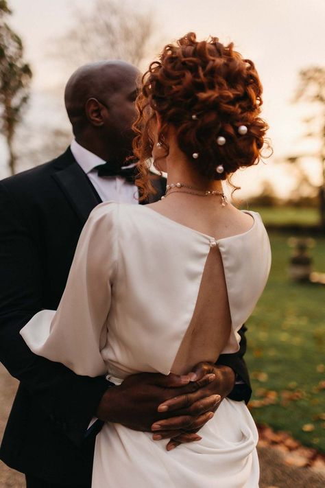 Groom in black tux with bowtie embracing bride in open back detail satin wedding dress with pearl hair accessories and pearl necklace Curly Updo With Pearls, Curly Hair Pearls, Boho Chic Hairstyles Wedding, Curly Updos For Wedding, Pearl Wedding Hairstyles, Wedding Updo Pearls, Bridal Updo Pearls, Shoulder Length Curly Wedding Hair, Pearl In Hair Hairstyles