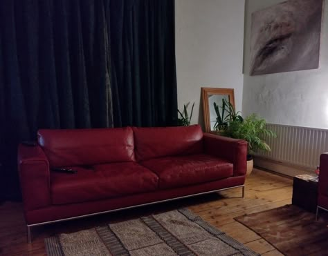 A long, low-backed, red leather sofa stands on a wooden floor. Dark green curtains behind. Abstract art to the right on the wall. A mirror and houseplants in the corner. A Turkish-style rug in the foreground. Red Sofa Aesthetic, Red Leather Sofa Living Room Ideas, Vintage Red Couch, Burgundy Leather Couch, Red Leather Couch Living Room, Red Leather Sofa Living Room, Red Leather Couch, Maroon Sofas, Red Leather Couches