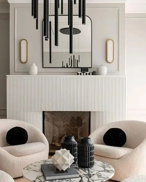 a contemporary space with a fireplace with a reeded surround, an arched mirror, creamy chairs, a black tube chandelier and black pillows Modern White Fireplace, Fluted Fireplace, Fireplace Trends, Modern Fireplace Mantel, Tube Chandelier, Minimalist Fireplace, Spec House, Mantel Surround, Fireplace Designs