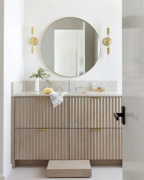 a greige fluted vanity with gold handles, a stone countertop, a round mirror, gold sconces and some lovely decor Fluted Bathroom Door, Fluted Vanity Bathroom, Greige Bathroom Vanity, Fluted Bathroom Vanity, Grey Terrazzo Floor, Greige Bathroom, Tiny Home Bathrooms, Fluted Vanity, Carved Sink