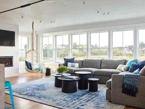 This room is all about the view, but in this small beach cottage, every inch counts. Designers decided to go with an extra long sectional (for the sitting space) and a fun hanging chair in the corner. The rest of the floor plan was kept open so that people can be sitting at the many turquoise stools at the island as well as the living space without additional furniture in between.  A fun assortment of various sized tables is an uncluttered look. See more at HGTV.com Corner Window Seats, Small Beach Cottage, Monochromatic Living Room, Wall Of Windows, Color Palette Living Room, White Cabinetry, Modern Cottage, Living And Dining Room, Beach Cottage